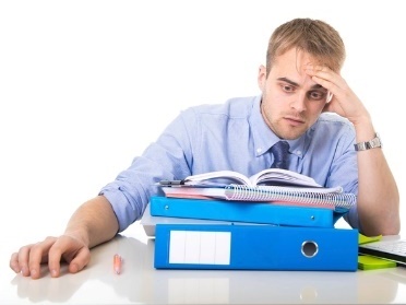 A person looking tired at a desk with a big stack of folders and documents in front of them.