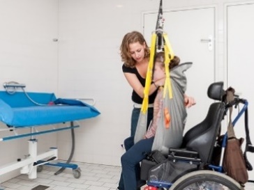 An NDIS worker using a hoist to help a participant out of their wheelchair and onto a changing table.