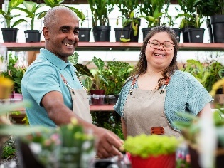 A participant and a support worker smiling in a garden nursery.