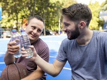 A participant and a support worker smiling and sitting on a basketball court together.