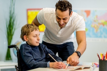A worker supporting a young particant write on a document in a classroom.