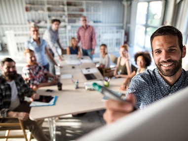 A group of people working together in a room. A person at the front of the room is writing on a whiteboard.