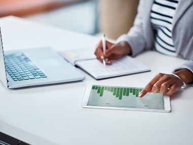 A person sitting at a desk writing notes. They are looking at data on a digital tablet and a laptop.