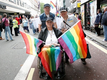 Two people smiling behind someone in a wheelchair, who is holding two rainbow flags. 